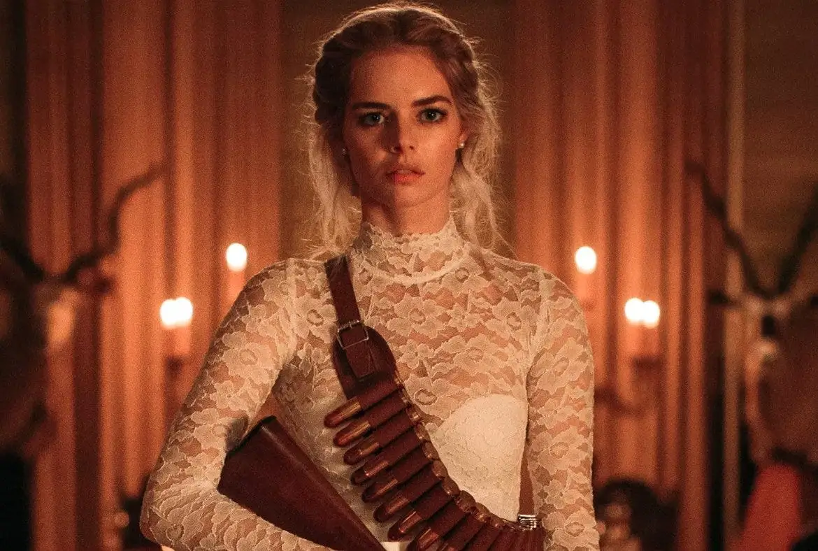 Samara Weaving as Grace in Ready or Not, standing in a candlelit room wearing a high-neck lace wedding dress with a shotgun strapped across her shoulder, her expression tense and alert amid the warm, ominous glow