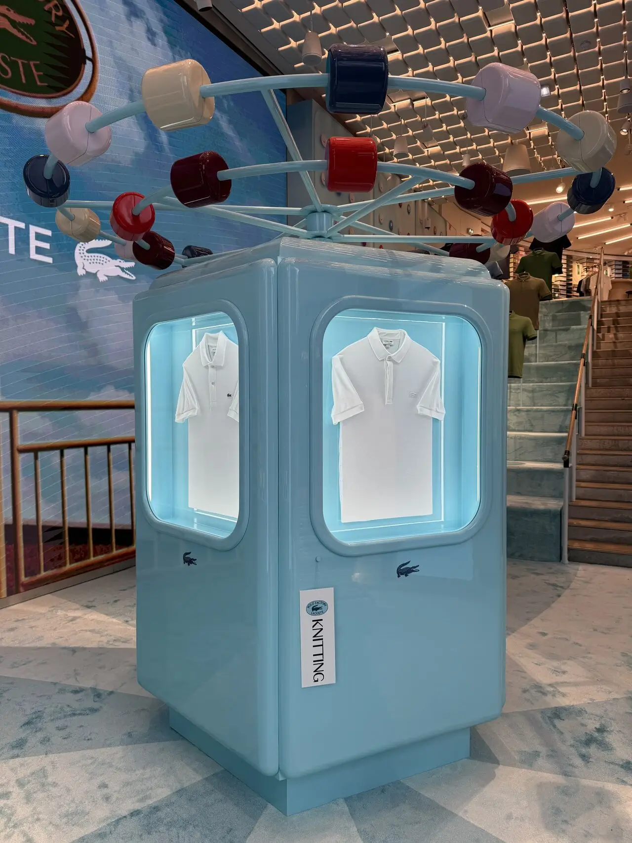 Lacoste Polo Factory Paris installation featuring illuminated polo shirts inside a pastel blue display unit labeled “Knitting,” surrounded by Atomic Age-inspired machinery and exhibition design