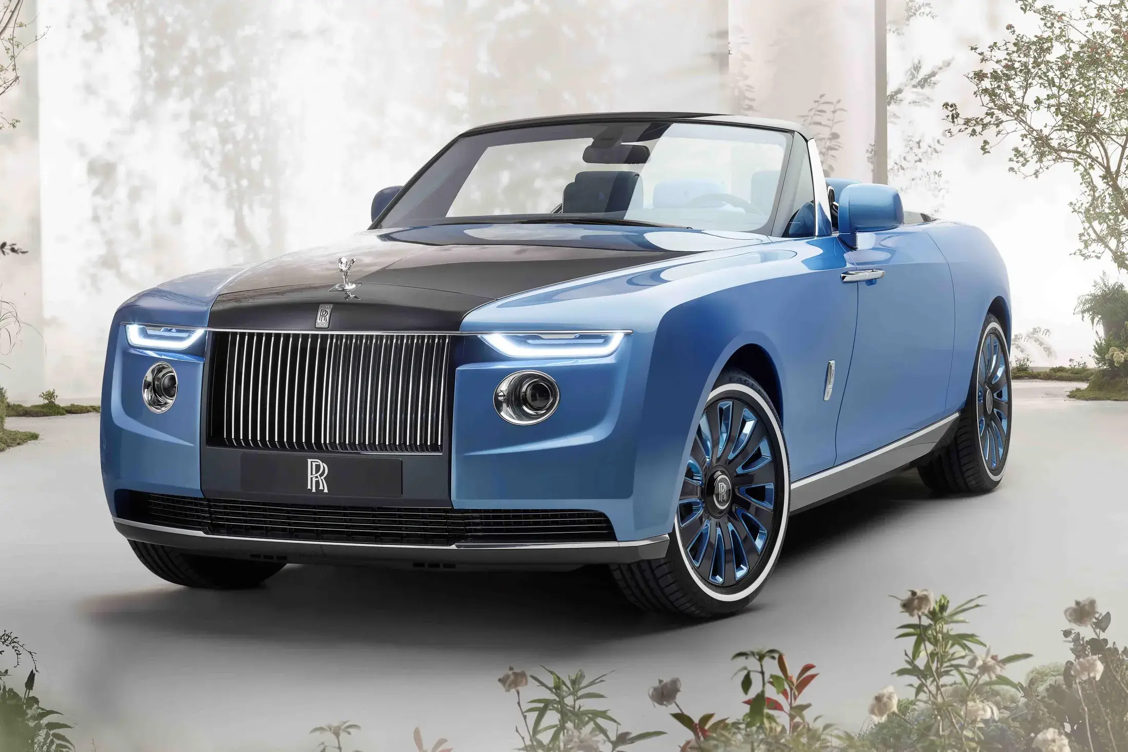 A bespoke Rolls-Royce Motor Cars coachbuilt convertible finished in a two-tone scheme—matte blue bodywork contrasted by a gloss black hood—featuring the marque’s signature Pantheon grille, illuminated LED headlamps, and Spirit of Ecstasy hood ornament. The car sits in a serene, garden-like setting with soft, diffused light, emphasizing its sculpted proportions, clean surfacing, and refined haute presence