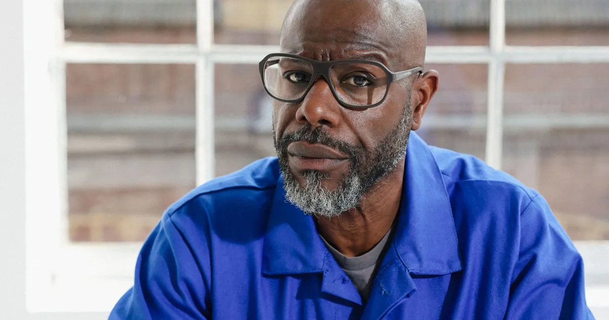 Portrait of Steve McQueen wearing glasses and a blue shirt, looking directly at the camera with a calm, contemplative expression against a softly lit interior background