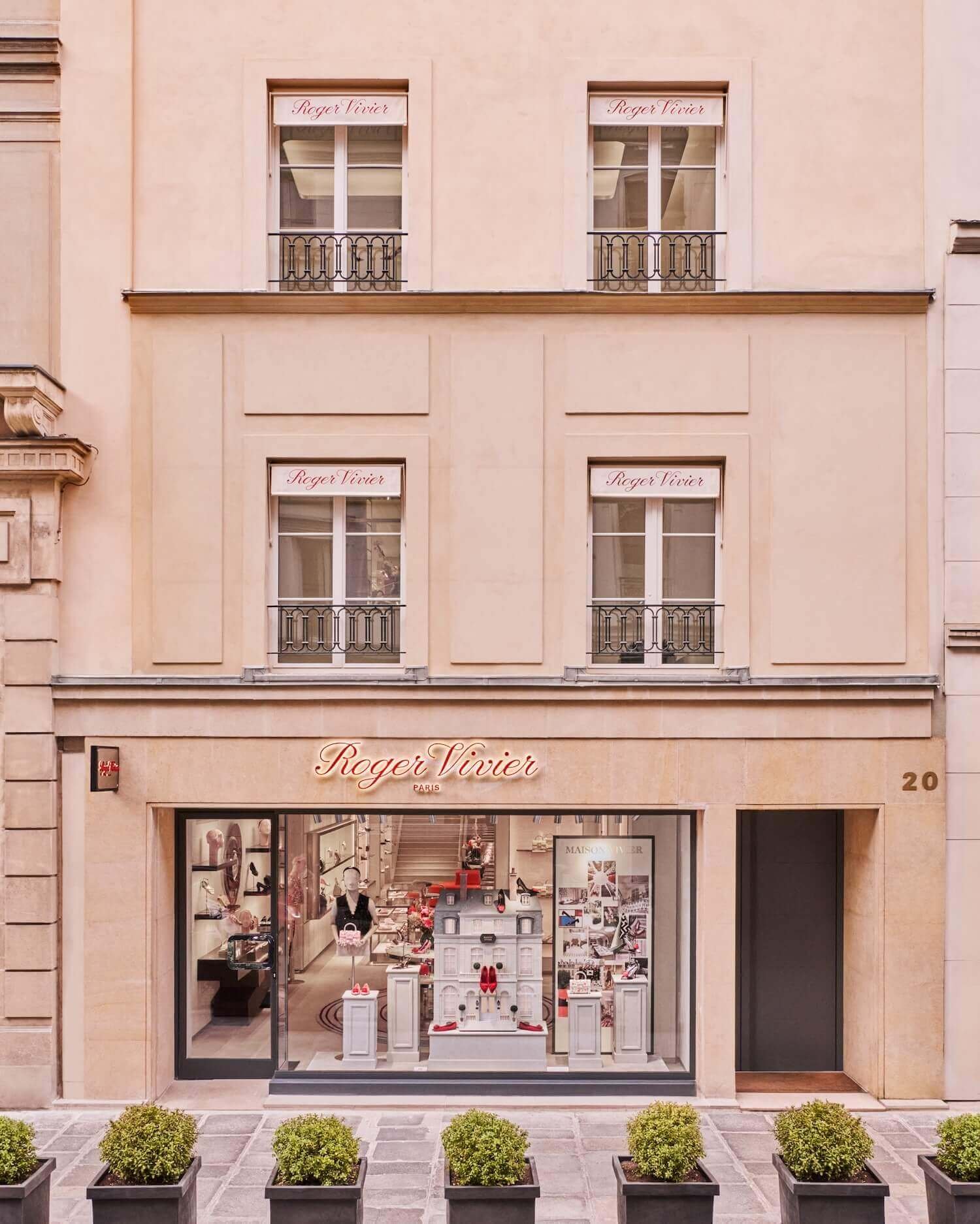 The exterior façade of the Roger Vivier Paris flagship on Rue du Faubourg Saint-Honoré presents a refined, sand-toned building with symmetrical windows framed by wrought-iron balconies. The boutique’s name is elegantly inscribed above the ground-floor display window, where curated installations of shoes and accessories are visible inside. Neatly aligned potted shrubs line the stone pavement, reinforcing the understated sophistication of the historic Parisian streetscape