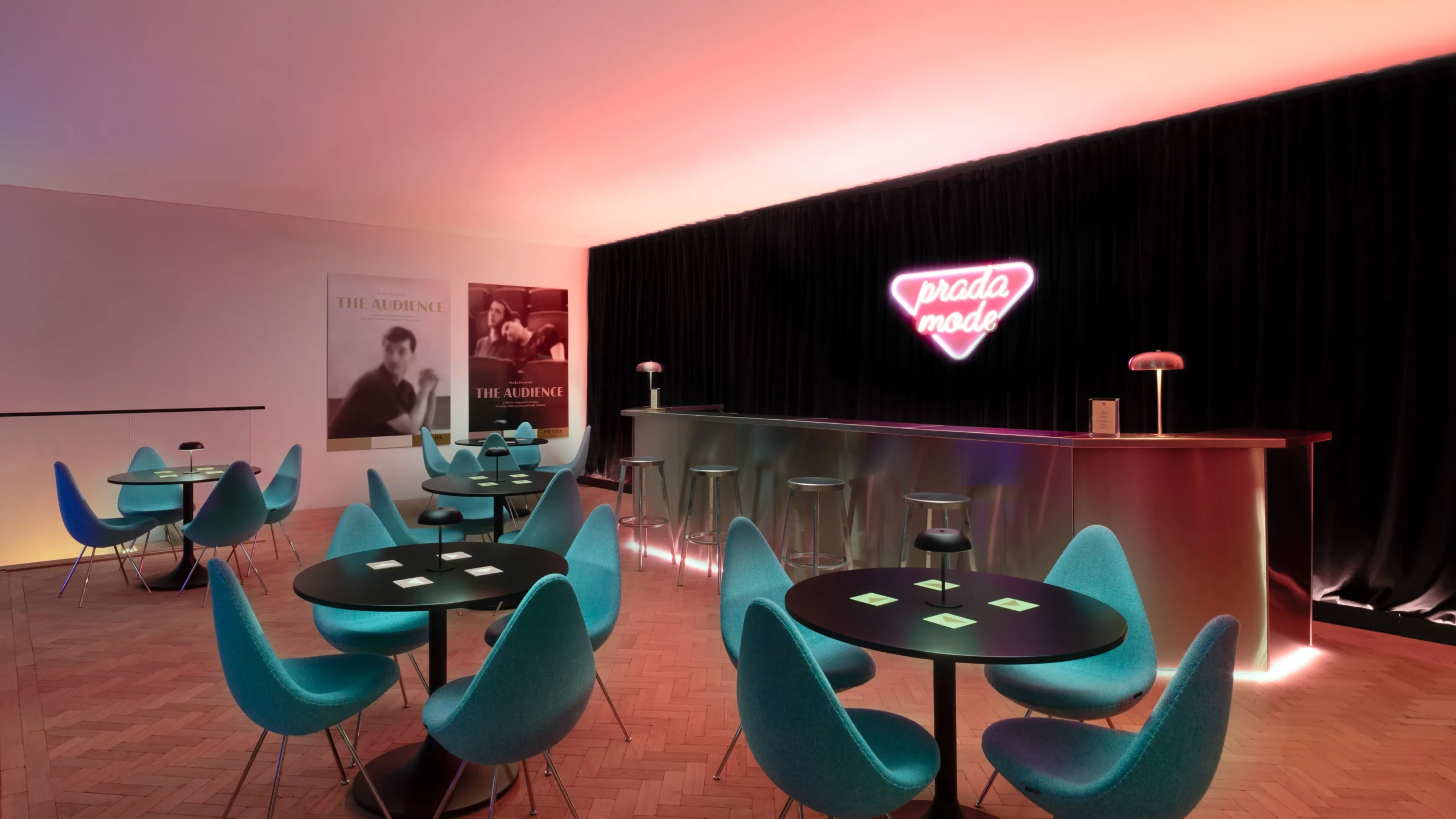 Prada Mode lounge installation featuring a neon “prada mode” sign glowing against black drapery, with a metallic bar, soft pink ambient lighting, and mid-century style teal chairs arranged around round tables in an intimate, cinematic interior setting