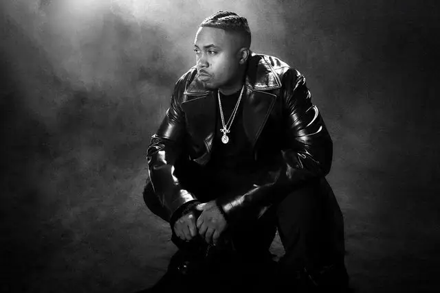 Black-and-white portrait of a man seated in a low, contemplative pose, wearing a leather jacket and chain necklace, lit dramatically from above against a dark, smoky studio backdrop, evoking a reflective and authoritative mood