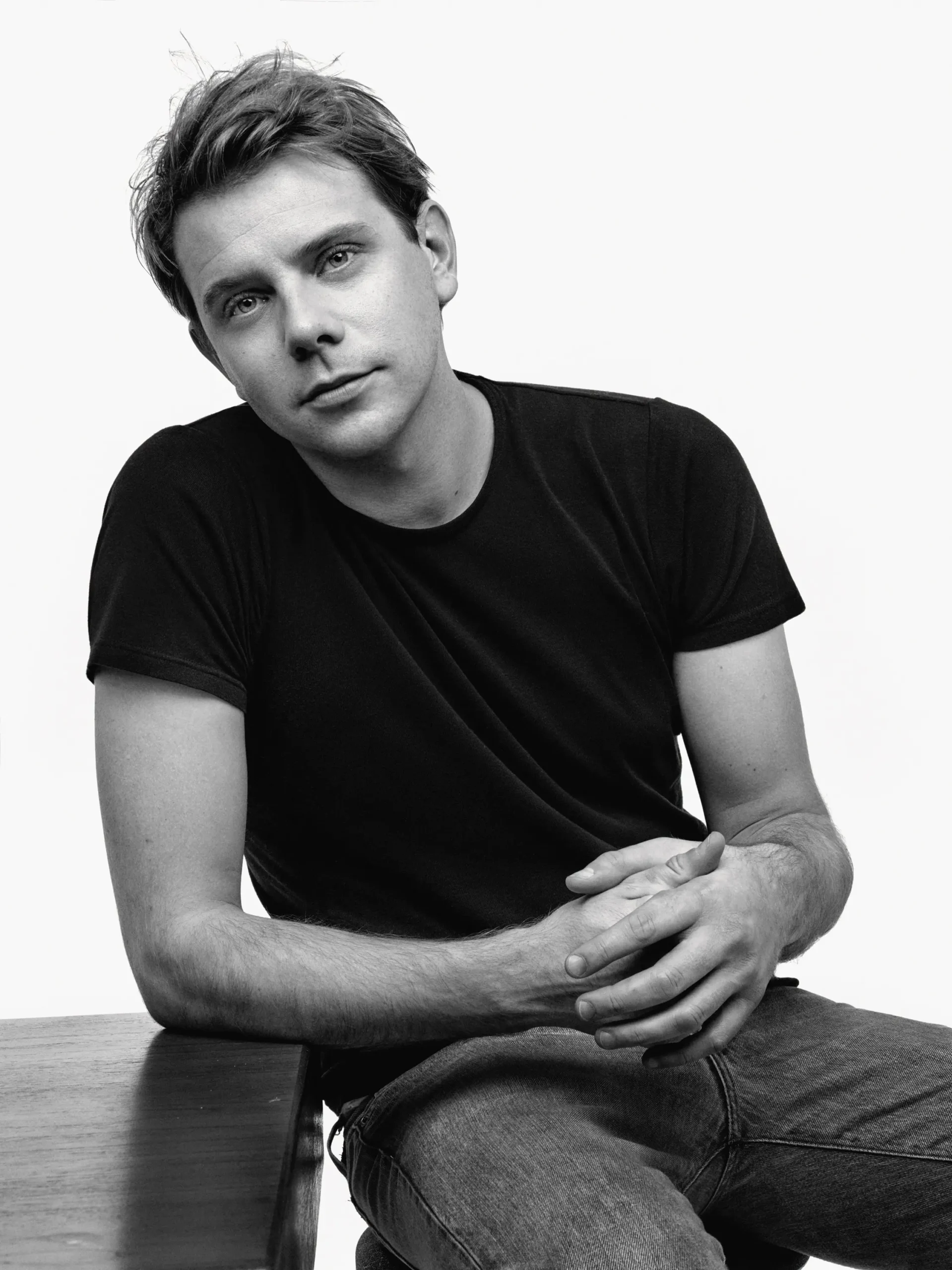 Black-and-white portrait of Jonathan Anderson seated casually in a plain t-shirt and jeans, hands clasped, reflecting a thoughtful and understated presence aligned with his design acknowledge