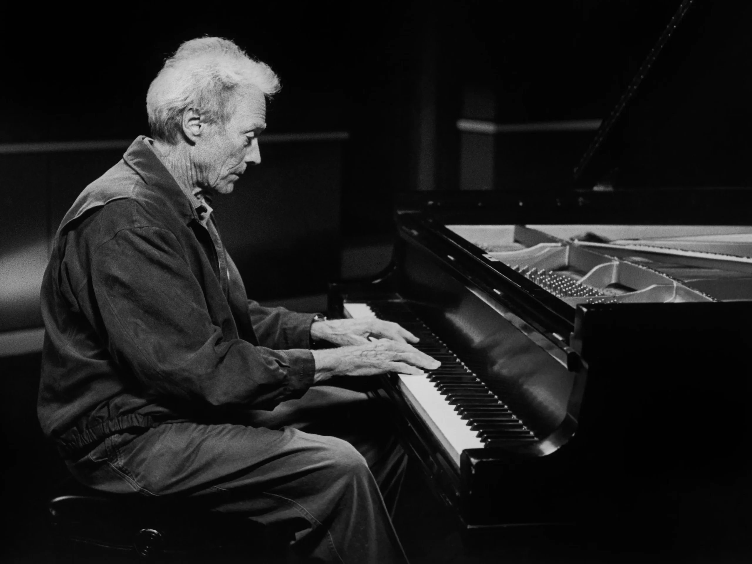 Clint Eastwood seated at a grand piano in a dimly lit space, captured in black and white as he plays with quiet concentration, emphasizing a reflective, late-life turn toward music and personal artistry