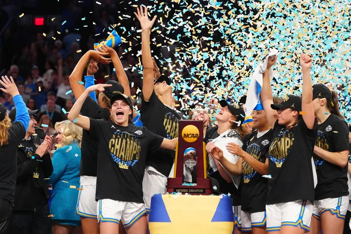 UCLA Bruins women’s basketball players celebrate around the NCAA championship trophy as confetti falls, marking their 79–51 victory over South Carolina in the 2026 title game