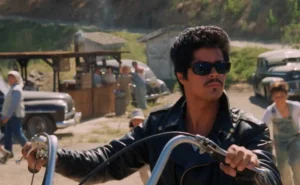 Bob Morales rides a motorcycle wearing a black leather jacket and sunglasses in a sunlit rural scene from La Bamba