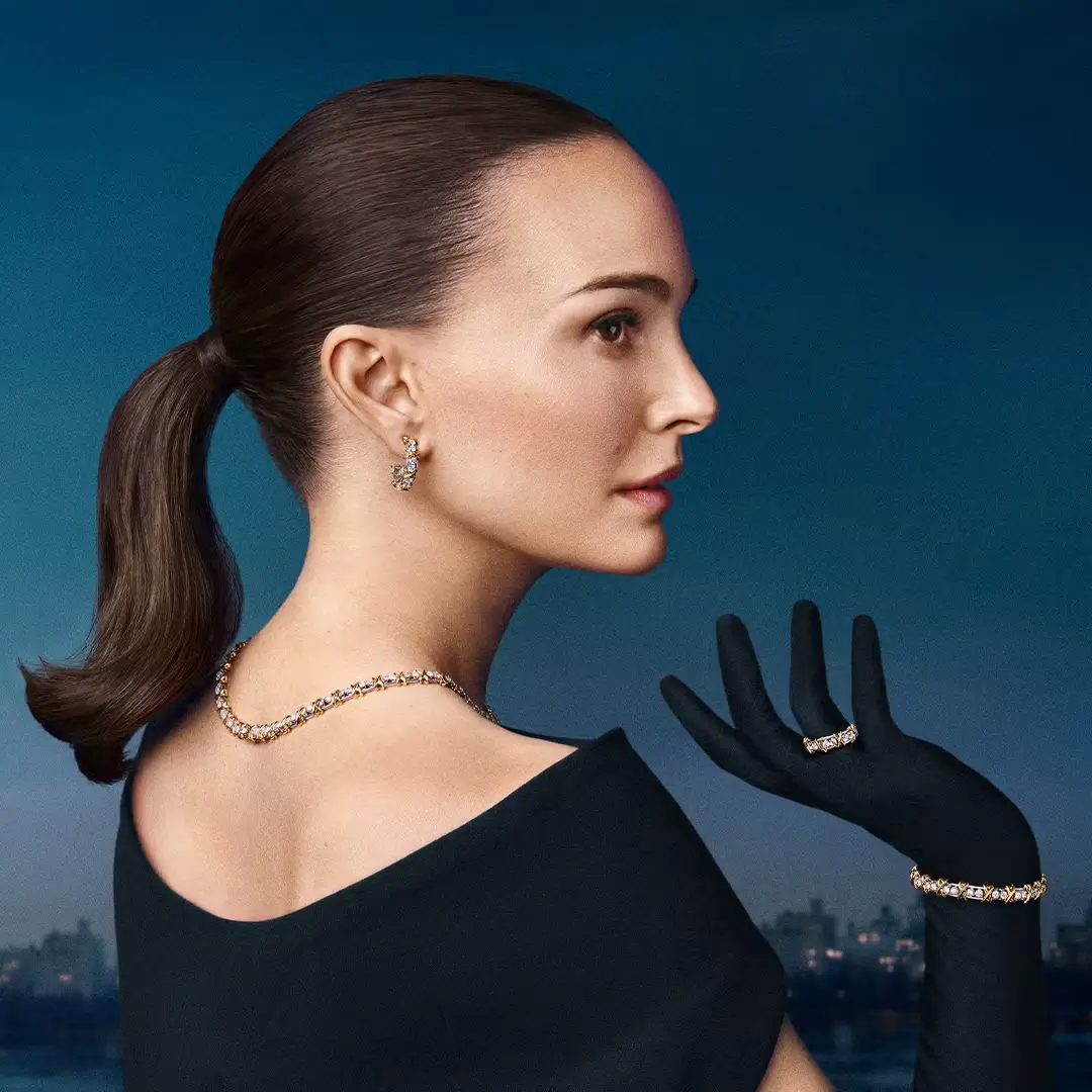 Natalie Portman in a Tiffany & Co. campaign wearing diamond necklace, earrings, bracelet, and ring against a deep blue evening skyline, styled in a black dress with a sleek ponytail and gloved hand gesture