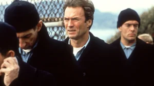 Clint Eastwood standing outdoors among a group of men in dark coats and knit caps, his expression focused and intense, captured in a cinematic still that evokes his iconic presence in gritty, character-driven roles