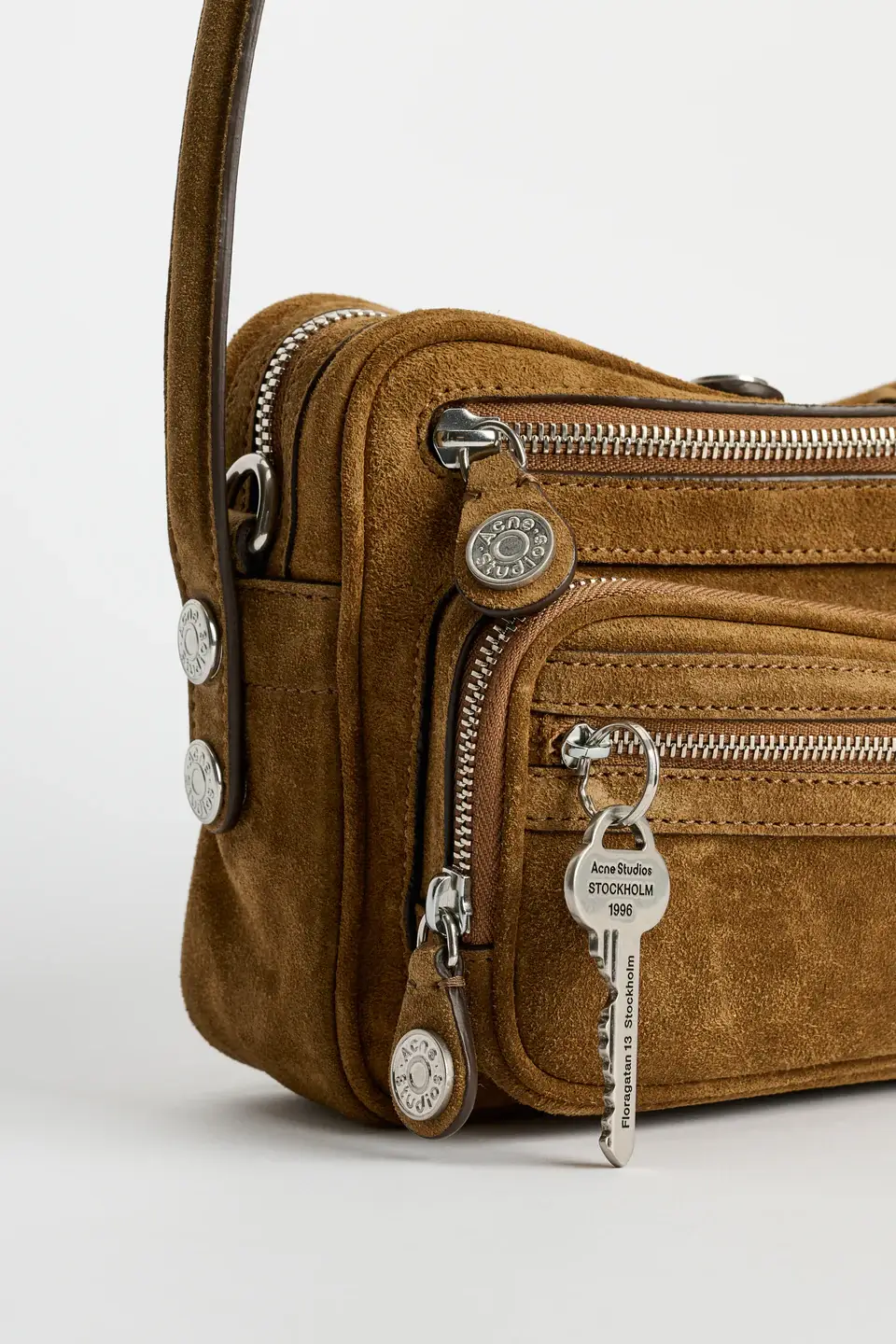 Close-up of Acne Studios Camero Party bag in cognac brown suede, highlighting layered zip compartments, silver-tone hardware, embossed zipper pulls, and signature key charm detail