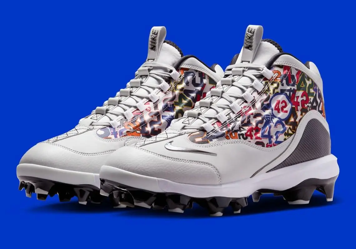 Three-quarter view of Nike baseball cleats in a white leather build featuring a multicolor collage of “42” graphics across the side panels, with white laces, a mini Swoosh, padded ankle collar, and black molded studs set against a vivid blue background