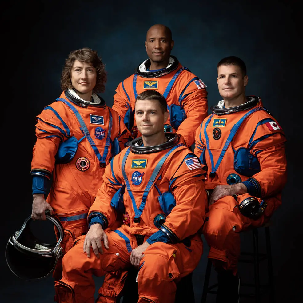 Four astronauts in bright orange flight suits pose for an official crew portrait against a dark studio backdrop, representing NASA’s Artemis II mission; two are seated in front while two stand behind, with mission patches, American and Canadian flags, and helmet gear