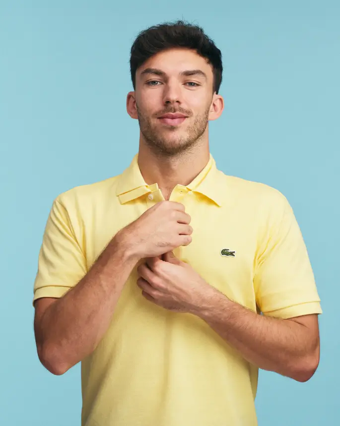 Portrait of a man wearing a pastel yellow Lacoste polo shirt, adjusting the collar, set against a clean light blue background with a minimal, modern aesthetic