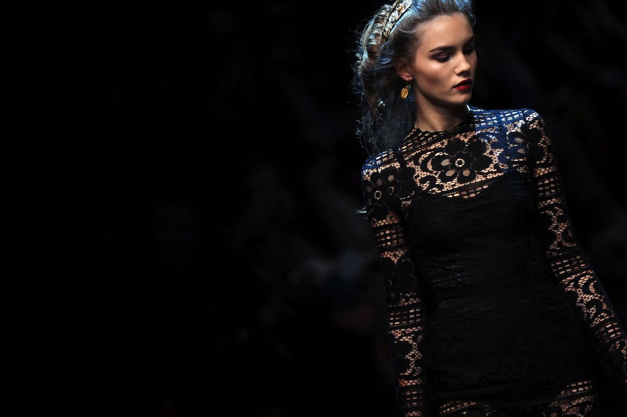 Model wearing a fitted black lace dress with intricate floral patterns, styled with a gold headband and earrings, walking under low, dramatic runway lighting against a dark background