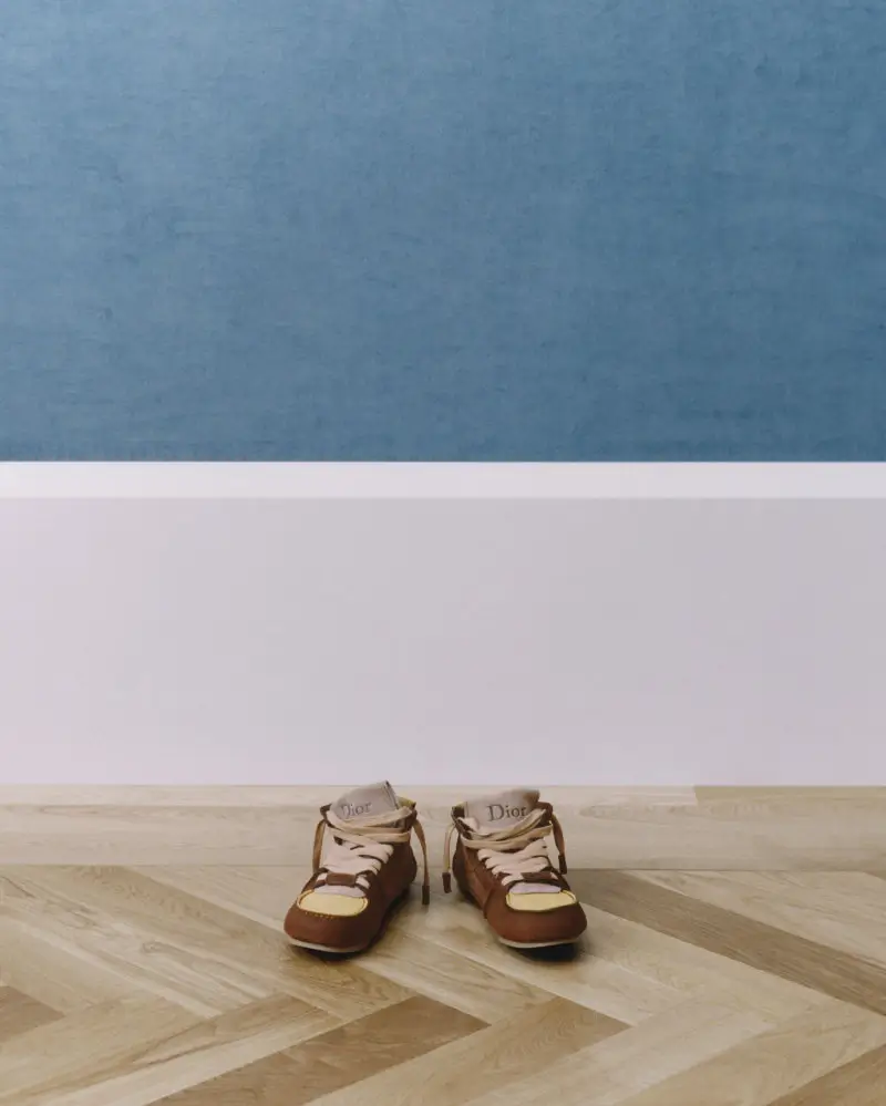 Dior Roadie shoes placed on a herringbone wooden floor, featuring brown leather construction, pale yellow suede toe panels, cream laces, and a minimal low-profile silhouette against a soft blue and white backdrop