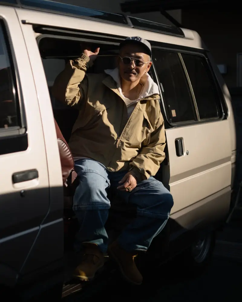 Man sitting in the open door of a van wearing a beige Timberland jacket, hoodie, relaxed-fit jeans, cap, sunglasses, and laceless Timberland boots with buckle closure in warm sunlight