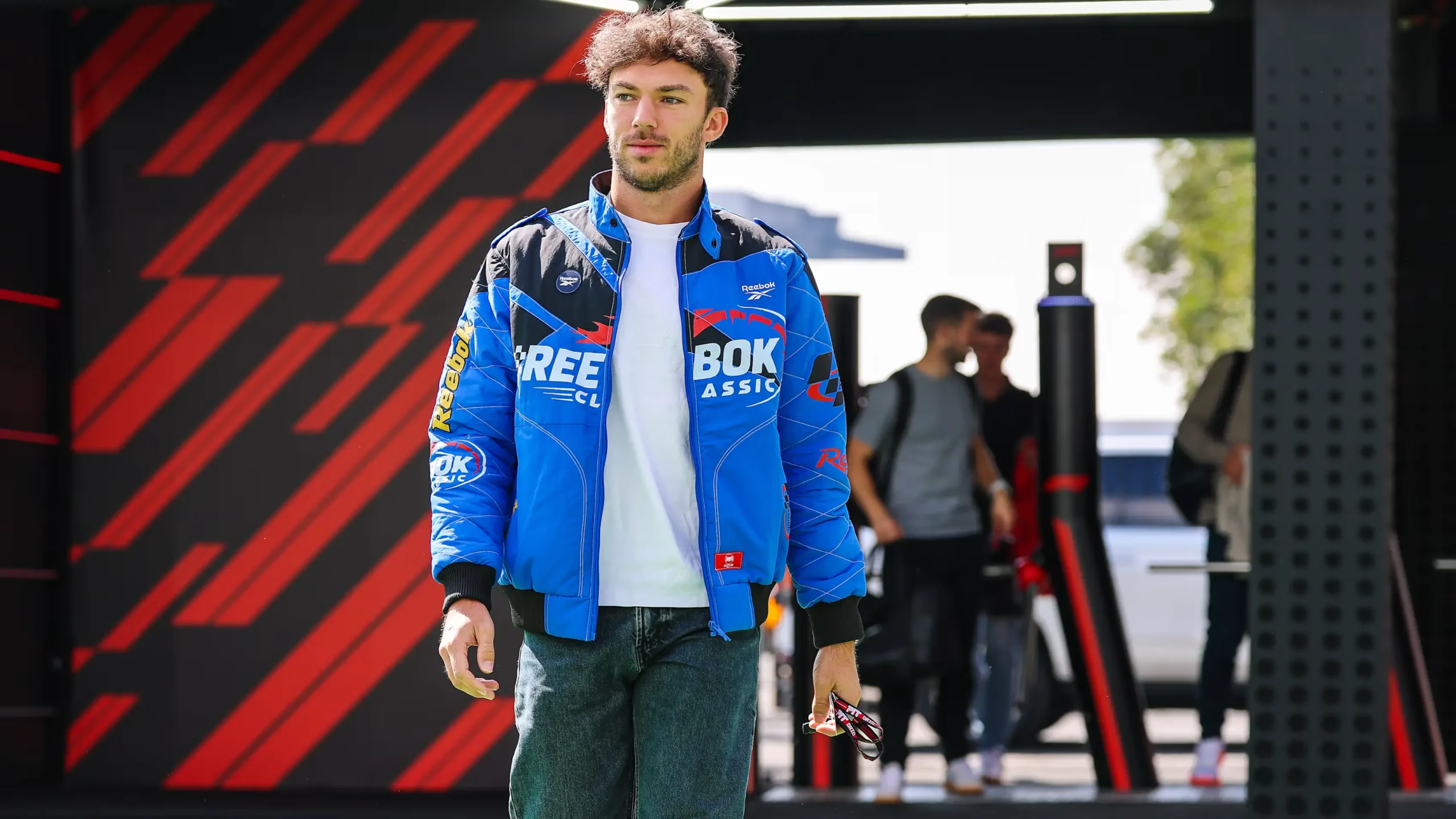 A man walking through a Formula 1 paddock area wearing a blue Reebok racing-style jacket over a white T-shirt and jeans, holding sunglasses, with a dynamic red-and-black graphic backdrop and blurred figures in the background