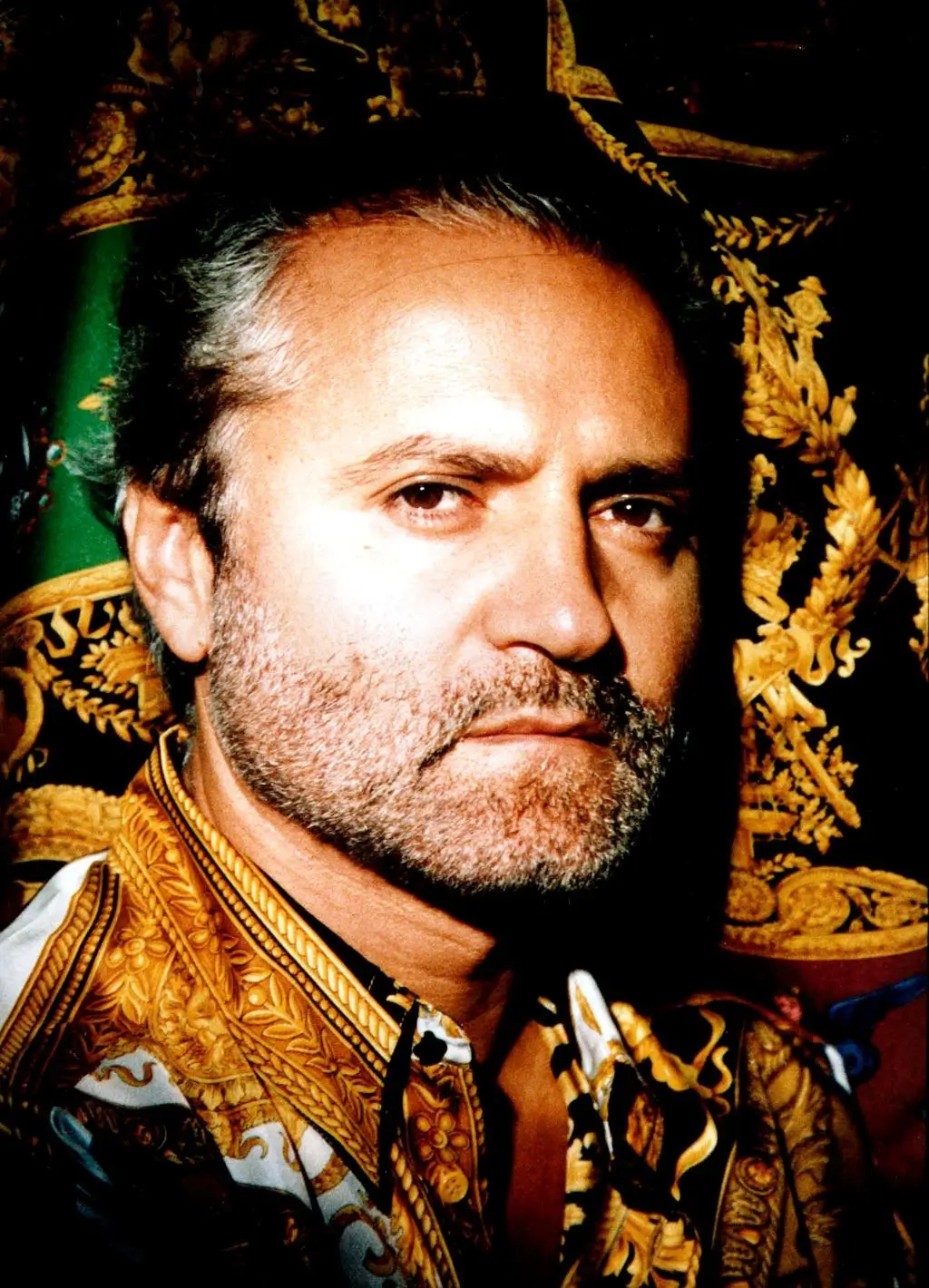 Portrait of Gianni Versace wearing a richly patterned baroque shirt with gold detailing, set against an ornate backdrop featuring classical motifs and deep black and green tones, emphasizing his signature aesthetic and view identity