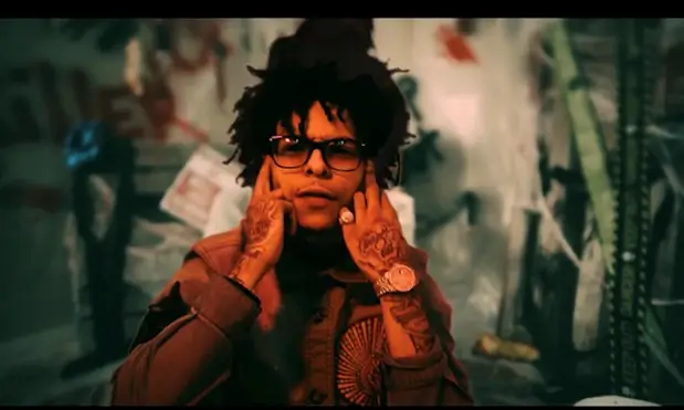 lil Tony performs in a dim, graffiti-covered setting, gesturing toward his face while wearing glasses and jewelry, capturing a moody West Coast music video aesthetic