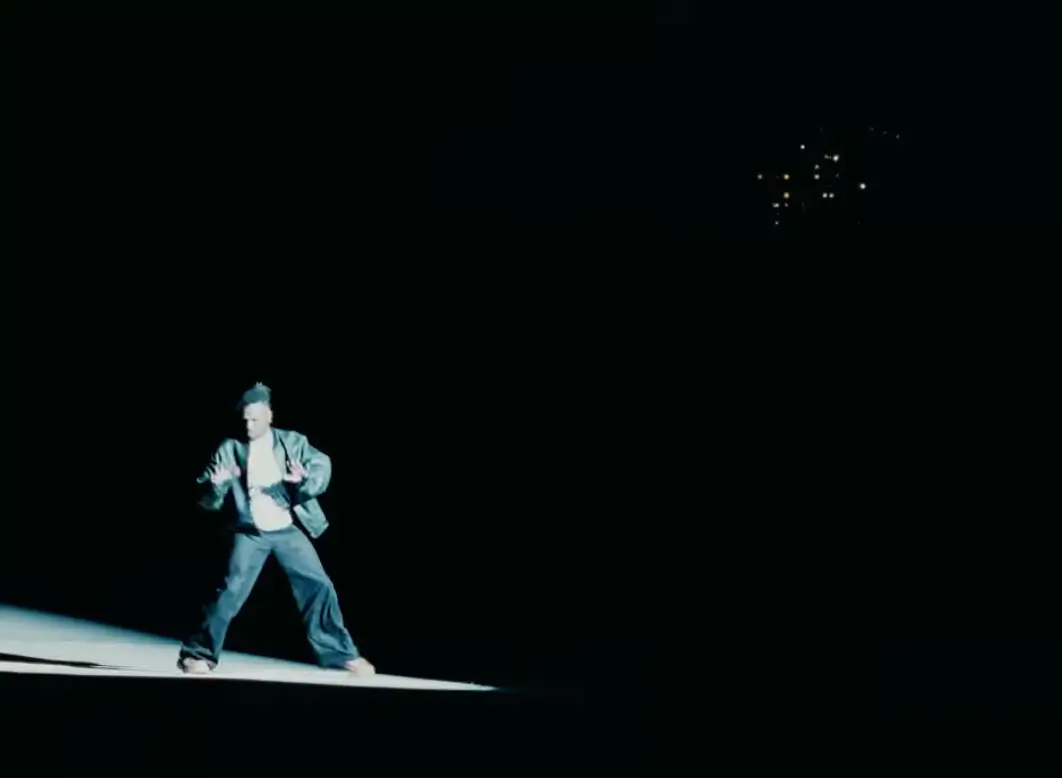 Isaiah Rashad stands under a harsh spotlight in a dark, empty space, body slightly crouched with hands raised mid-motion, surrounded by deep black negative space that isolates the subject and heightens a restrained, introspective mood