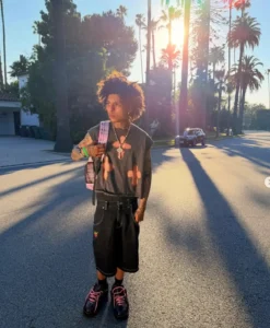 lil Tony stands in the middle of a sunlit Los Angeles street lined with palm trees, wearing casual streetwear and a backpack, capturing a laid-back West Coast aesthetic