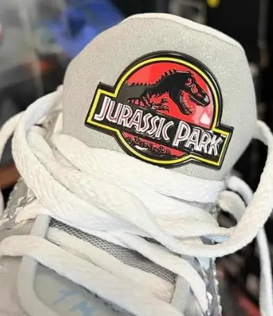 Close-up of the tongue on a metallic silver Nike Ja 3 shoe featuring a stitched Jurassic Park logo patch in red, yellow, and black, with white laces layered across the front