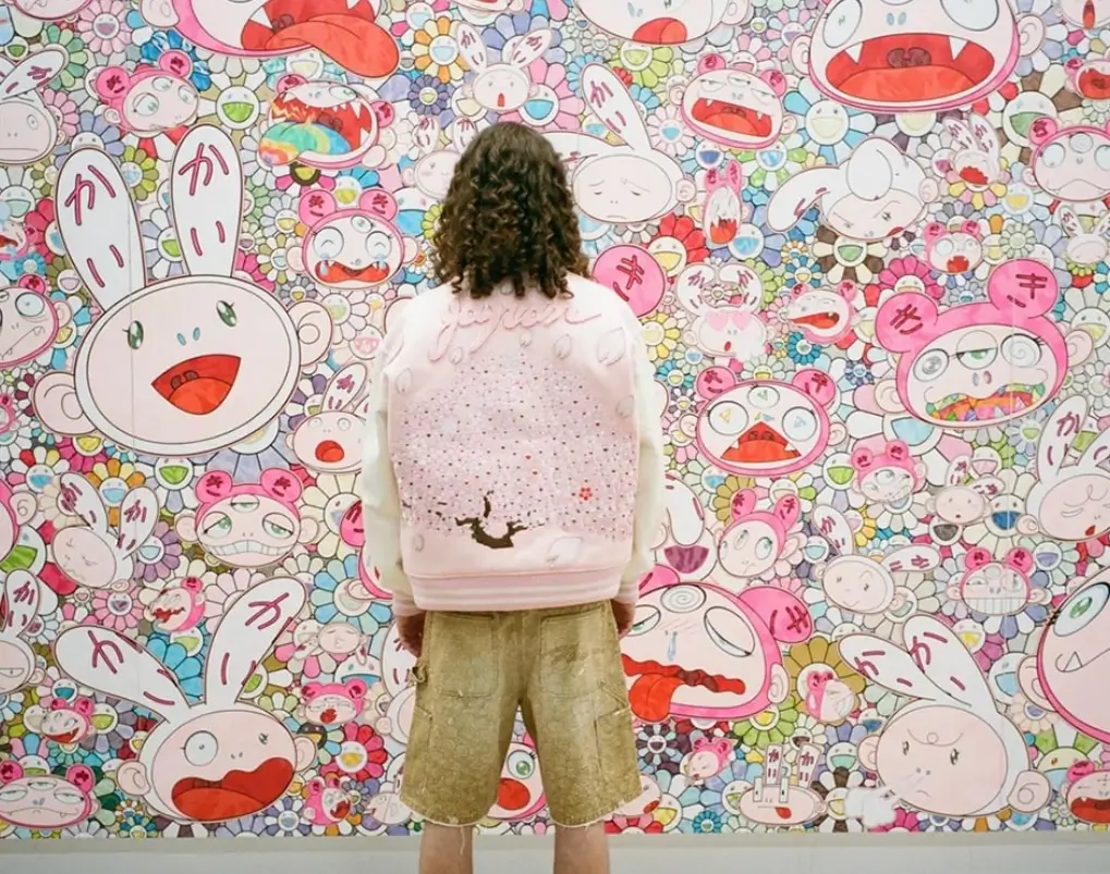Visitor wearing a pale pink jacket stands facing a large-scale Takashi Murakami-style mural filled with colorful cartoon characters, smiling flowers, and surreal faces, blending pop art and Japanese superflat aesthetics in a dense, immersive composition