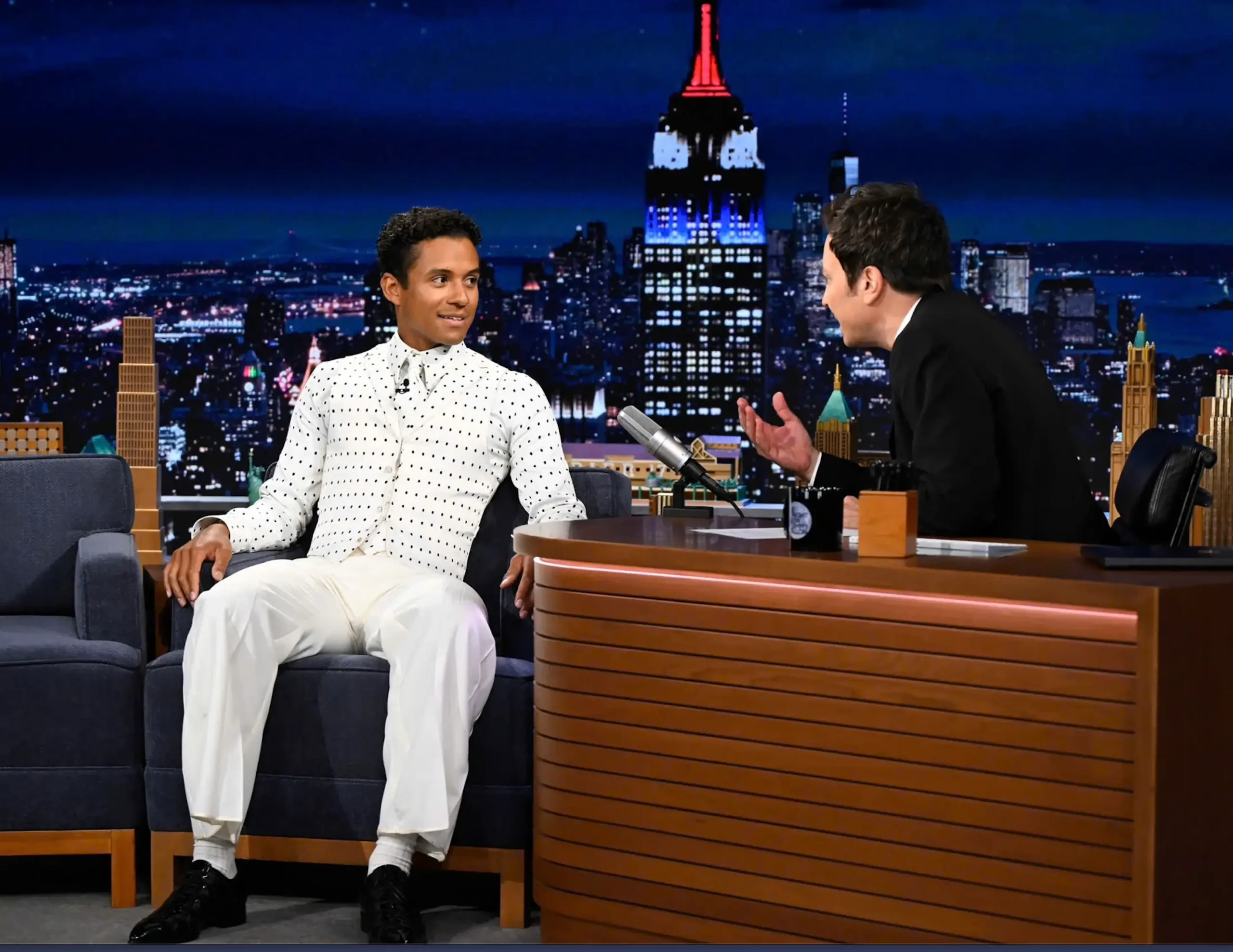 Jaafar Jackson seated on The Tonight Show couch wearing an all-white polka-dot suit, speaking with host Jimmy Fallon against a New York City skyline backdrop