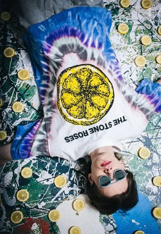 SAINT Mxxxxxx tie-dye T-shirt featuring The Stone Roses lemon graphic worn by model lying on splatter-painted surface with scattered lemon slices