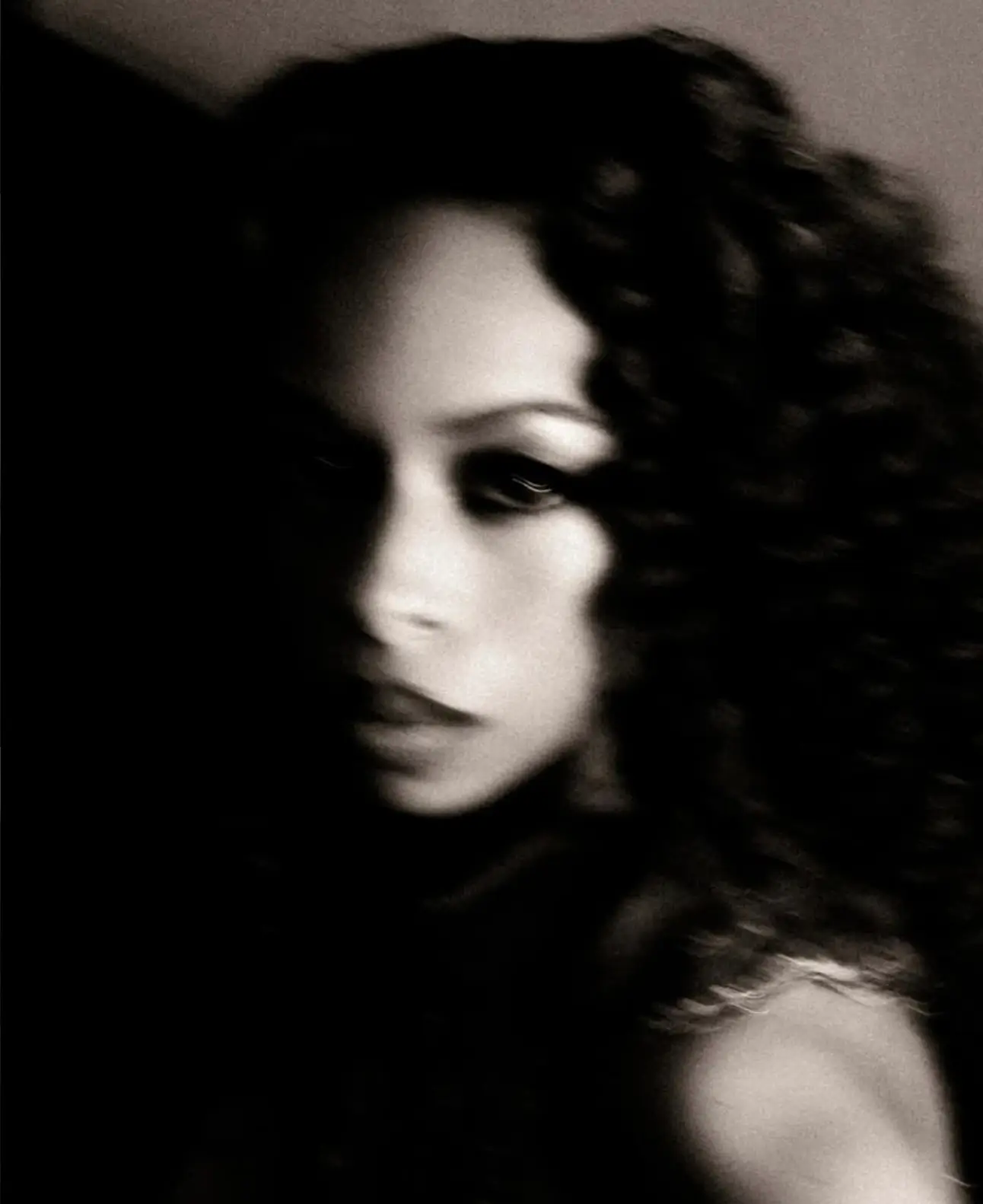 Soft-focus black-and-white portrait of a woman with voluminous curly hair, partially obscured in shadow, featuring blurred contours and a moody, atmospheric expression