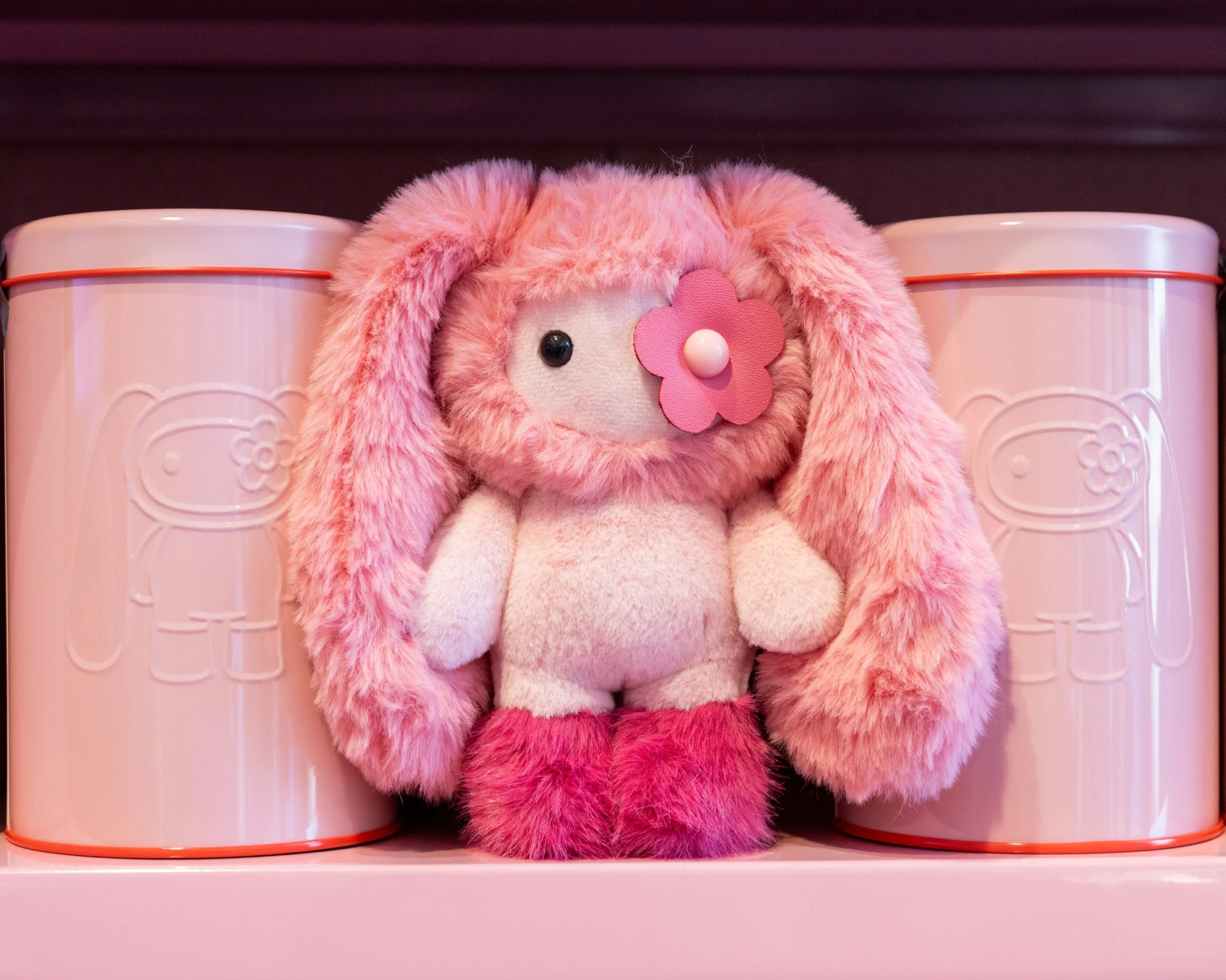 Pink juju plush collectible by Cj Hendry displayed between branded storage tins, highlighting soft texture, character design, and collectible art toy presentation at Phillips Hong Kong exhibition