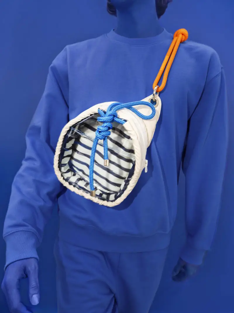 Model wearing a Jack Gomme crossbody bag with a cylindrical silhouette, featuring a transparent striped body, cream padded trim, and blue drawcord closure, suspended from a bright orange rope strap against a monochromatic blue backdrop