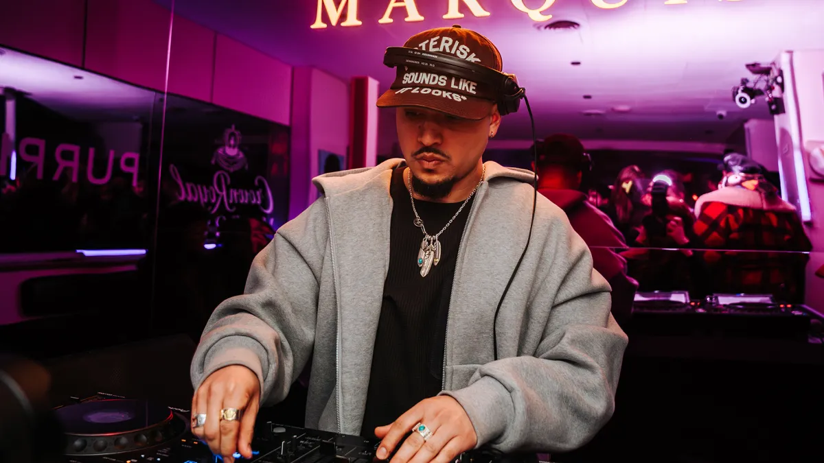 Joe Kay DJing live behind the decks in a dimly lit venue, wearing a black cap and grey hoodie, adjusting controls on a mixer under pink ambient lighting as a crowd reflects in the background, capturing the intimate energy of a Soulection set