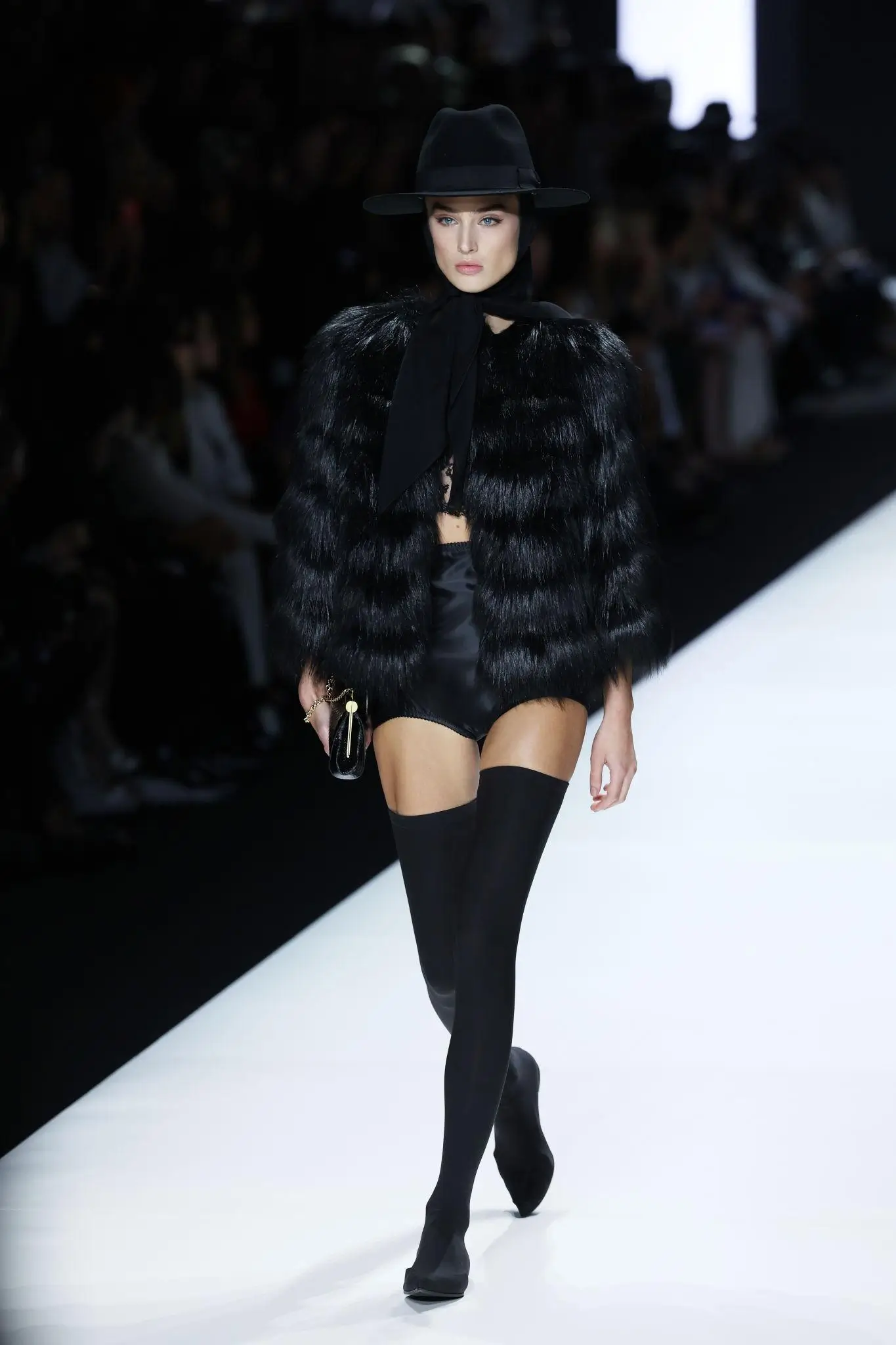 Model walking a runway in an all-black look featuring a wide-brim hat, a textured fur jacket, a high-cut bodysuit, and thigh-high stockings, accessorized with a small clutch, set against a bright catwalk with a blurred audience in the background