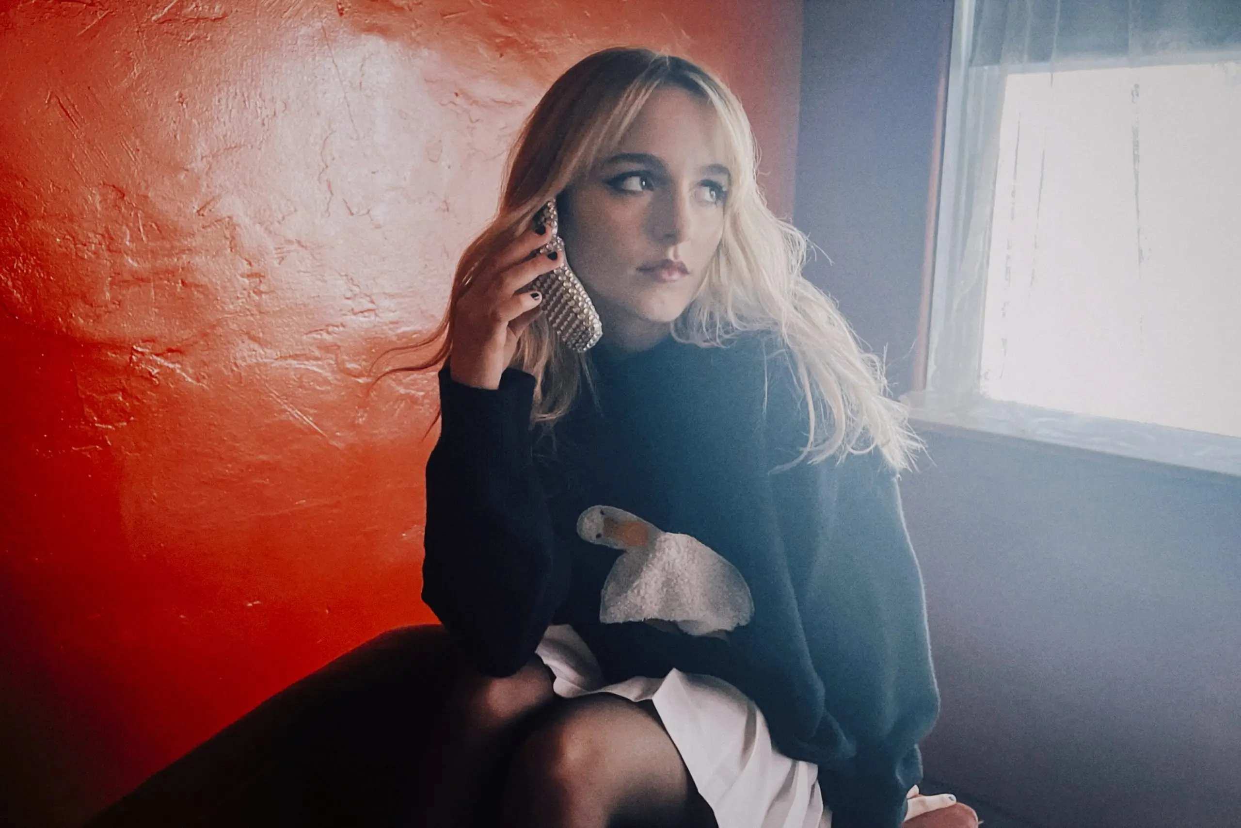 Mckenna Grace seated by a window in soft natural light, holding a rhinestone-covered phone to her ear, wearing a black sweater with a swan motif against a textured red wall backdrop