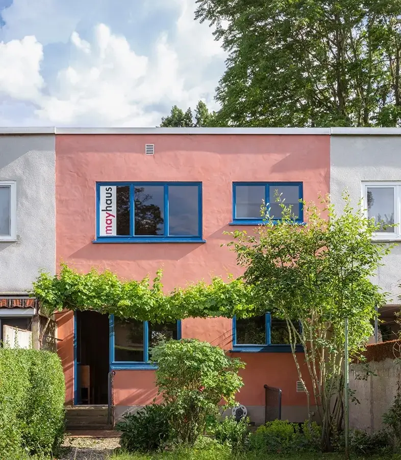 Restored Mayhaus in Frankfurt featuring a pastel pink modernist facade, flat roof, blue-framed windows, vine-covered entry, and landscaped garden with trees and shrubs