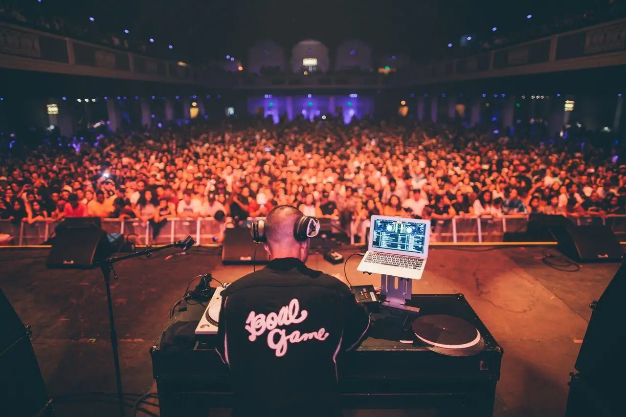 Overhead view of a DJ setup featuring a laptop and mixer on a small table, positioned on a dimly lit stage with warm lighting and speakers in the background, capturing the behind-the-scenes perspective of a live music performance environment