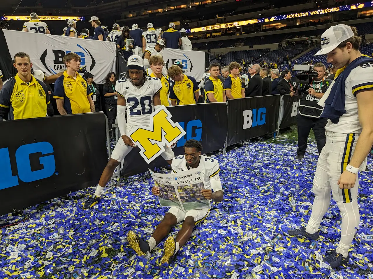 Michigan Wolverines players celebrate on a confetti-covered court after winning the Big Ten championship, with one player holding a block “M” logo while another reads a newspaper