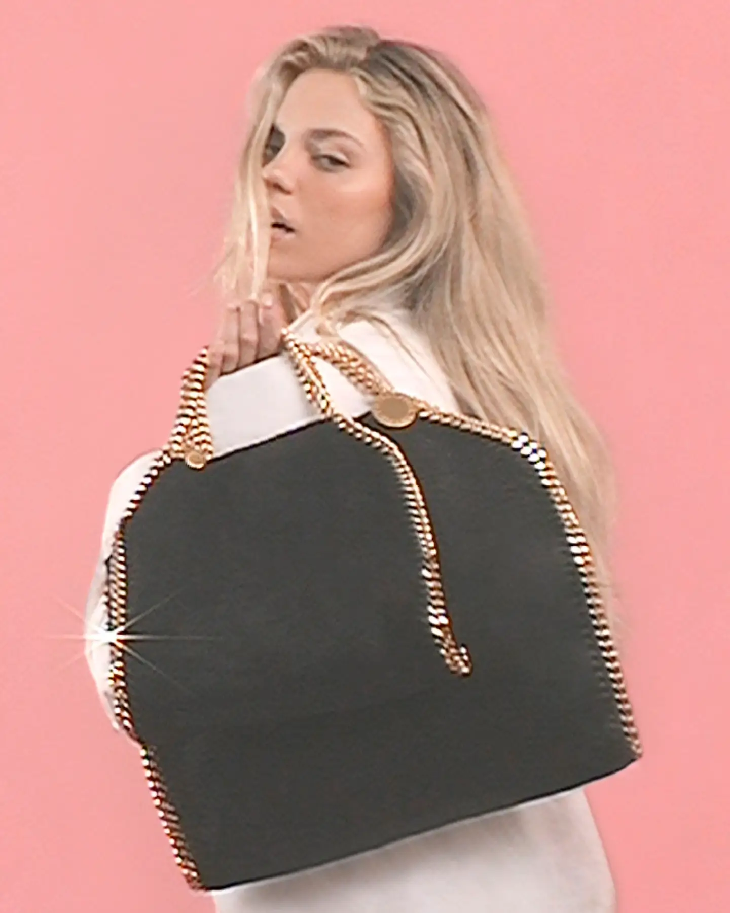 Renee Rapp turns slightly over her shoulder against a soft pink backdrop, holding a large black Stella McCartney Falabella tote with signature gold chain trim and handles, the bag’s structured yet slouchy silhouette catching light along its metallic edges