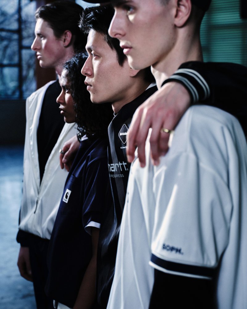 Group of models in Carhartt WIP x F.C.Real Bristol apparel stand shoulder-to-shoulder in profile, wearing coordinated football-inspired jerseys and jackets in black, navy, and white, highlighting co-branded details in a clean, editorial setting