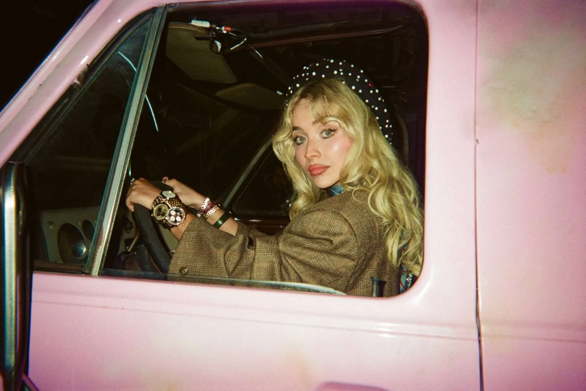 Sabrina Carpenter sits in the driver’s seat of a pink vintage car, looking back toward the camera while wearing a polka-dot headscarf and layered jewelry