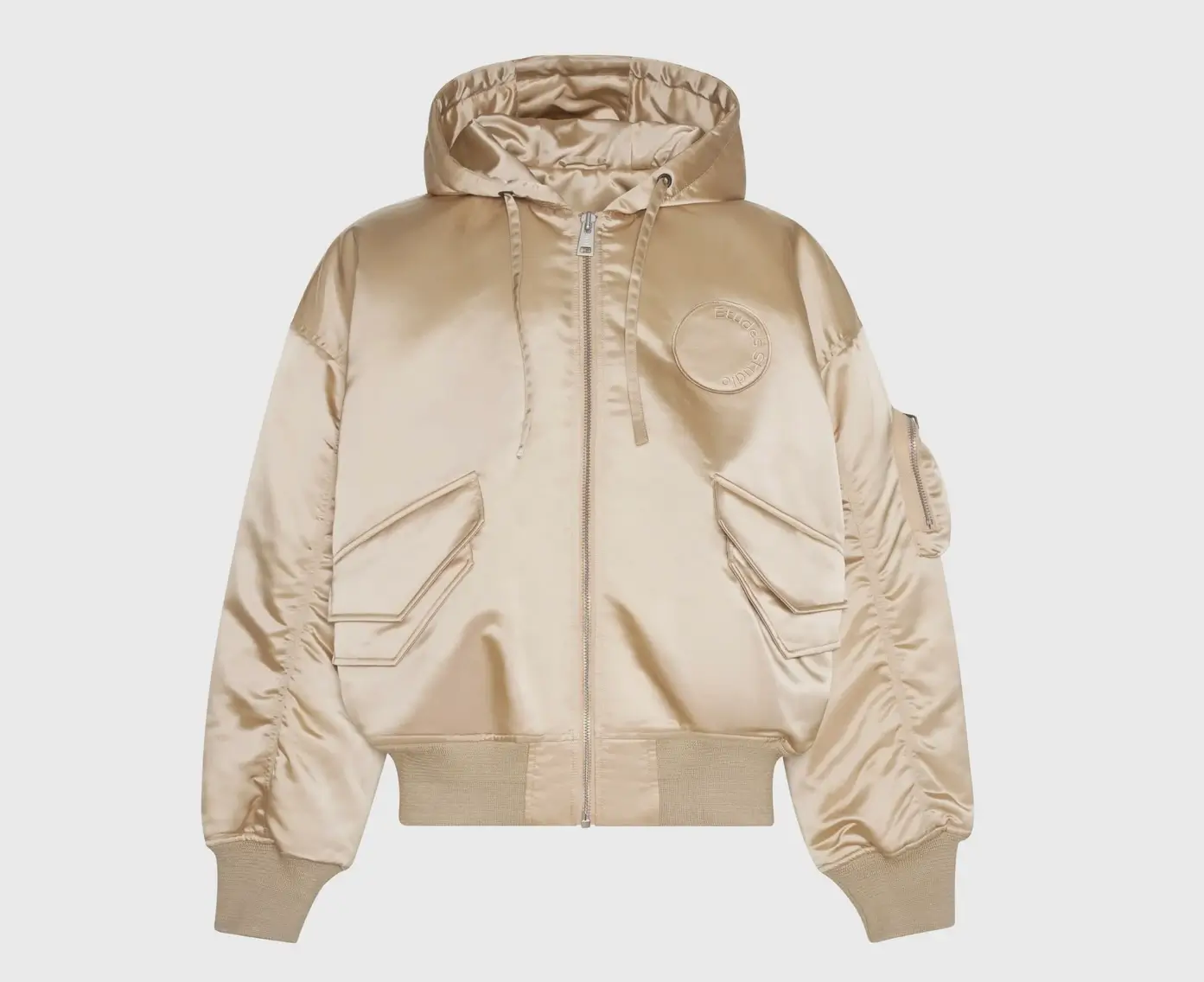 Champagne gold oversized nylon bomber jacket with hood, featuring front zip closure, ribbed cuffs and hem, angular flap pockets, sleeve utility pocket, and embossed circular logo patch on chest