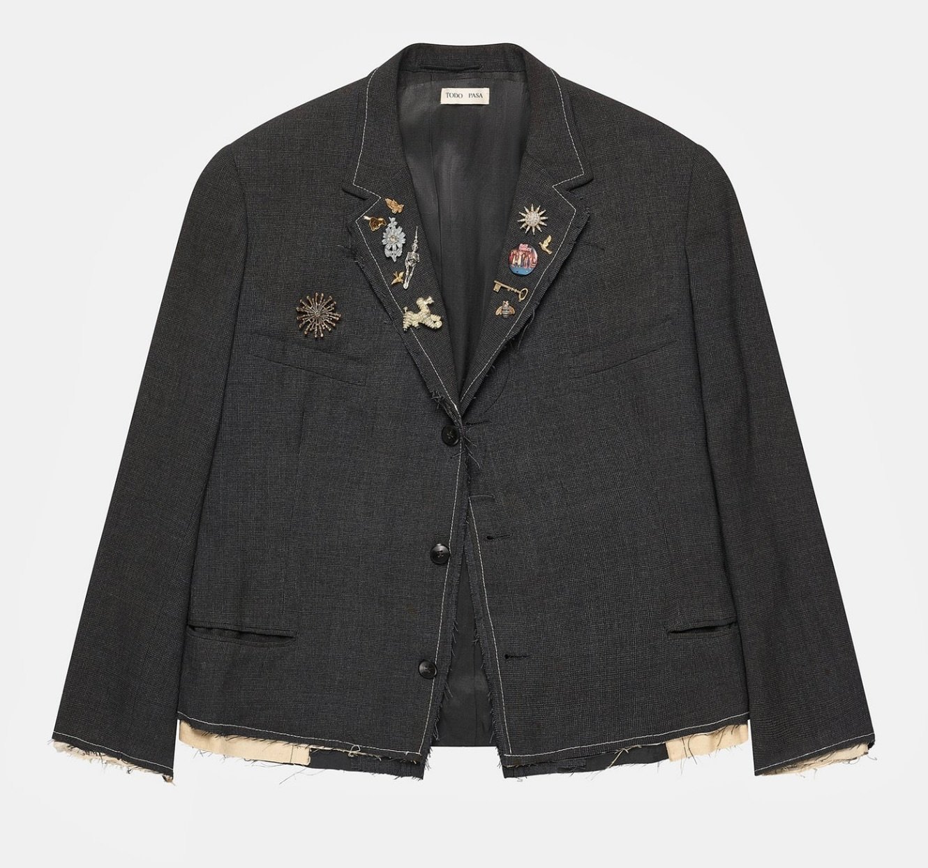 Todo Pasa “Sol” Clavo Jacket in dark textured wool featuring raw unfinished hems, contrast stitching, and an array of eclectic pins including a sun motif, key, and ornamental charms across the lapel
