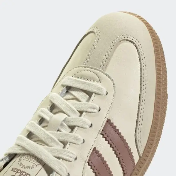Close-up of cream Adidas shoe upper with tonal stitching, soft leather panels, matching laces, muted pink Three Stripes, and a gum sole edge