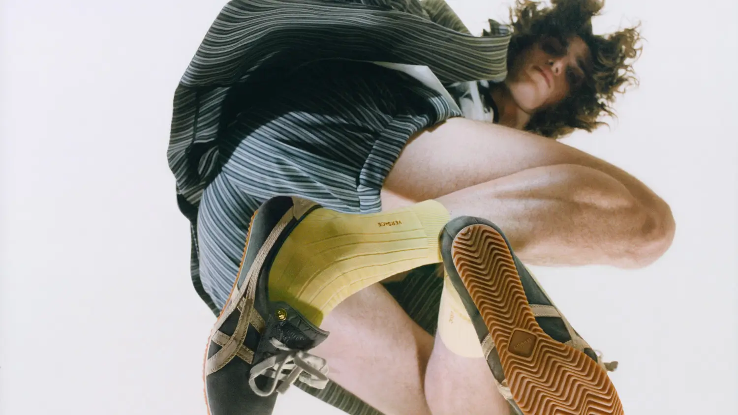 Low-angle campaign shot of a model in a striped outfit wearing Onitsuka Tiger x Versace Tai-Chi Sakura shoe emphasizing the gum outsole, curved low-profile silhouette, and subtle metallic detailing against a minimal sky backdrop