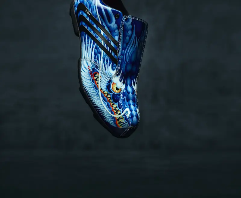 Floating angled view of a blue Y-3 F50 TUNIT boot with intricate dragon artwork, fold-over tongue, and black adidas stripes against a dark gradient background