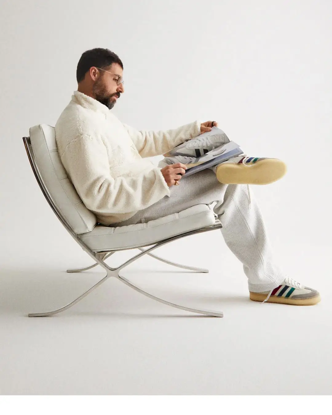 Side-profile portrait of Ronnie Fieg seated in a minimalist modern chair, wearing a cream fleece and grey sweatpants, reading a book while showcasing neutral-toned adidas sneakers with gum soles against a clean white studio background