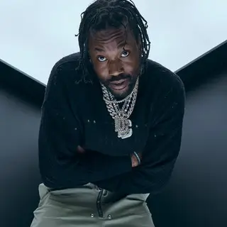 Portrait of Meek Mill leaning forward with arms crossed, wearing layered diamond chains and a black sweater against a minimal light background, looking directly at the camera