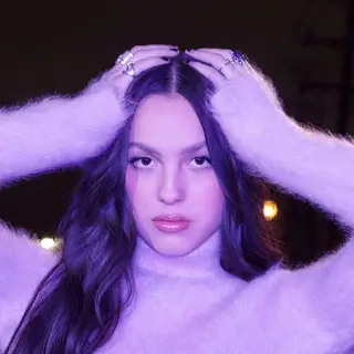 Olivia Rodrigo in a soft purple-lit portrait, wearing a fuzzy sweater with hands placed on her head, conveying an intimate and emotionally introspective mood