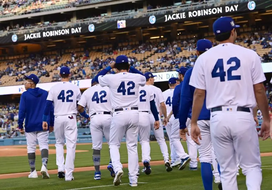 Players from a Major League Baseball team walk onto the field wearing white uniforms with the number 42 during Jackie Robinson Day, with stadium signage displaying the tribute and fans visible in the stands